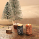 Bougies artisanales en cire naturelle avec motif de cerf, allumées sur une table en bois avec sapins enneigés en arrière-plan, créant une ambiance hivernale chaleureuse.