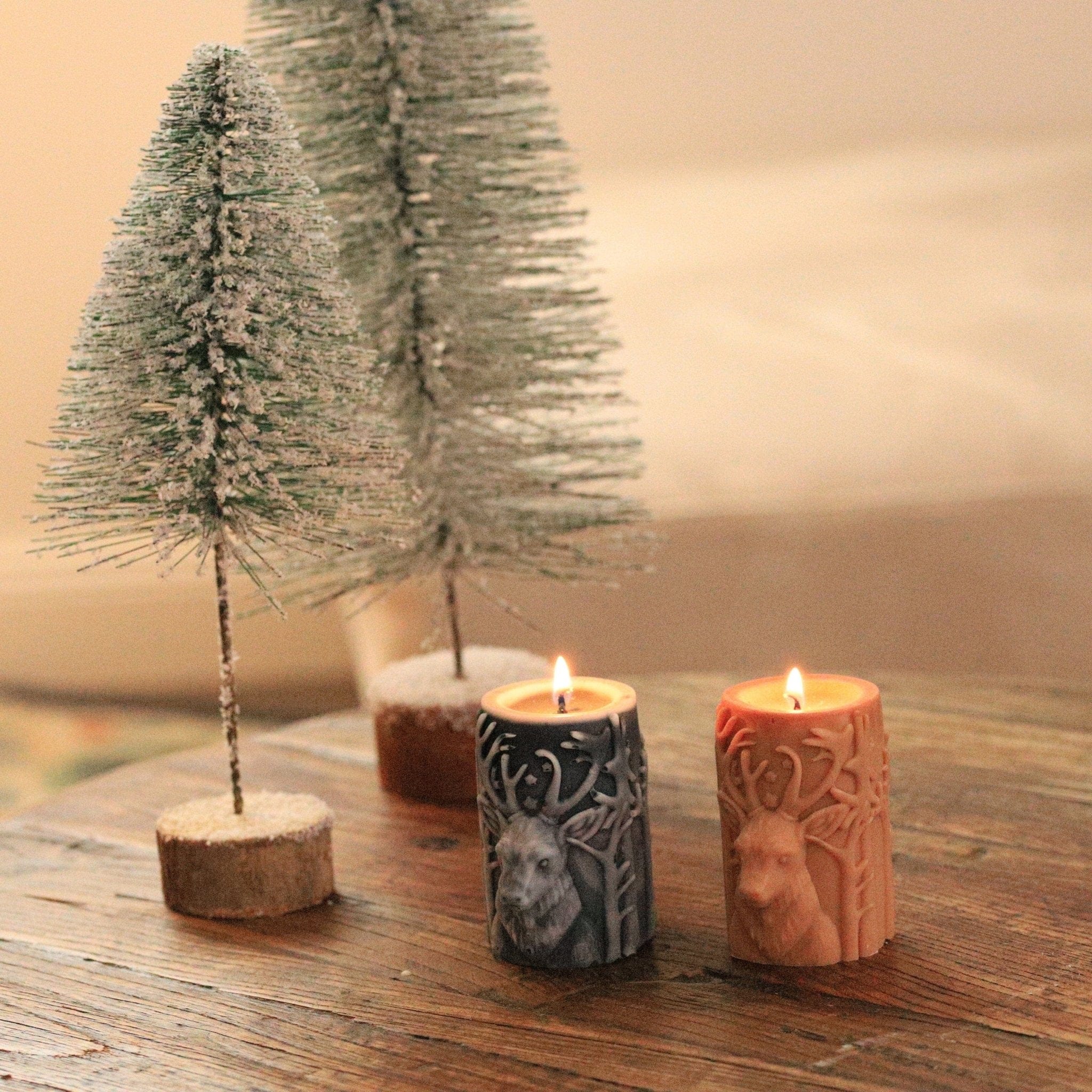 Bougies artisanales en cire naturelle avec motif de cerf, allumées sur une table en bois avec sapins enneigés en arrière-plan, créant une ambiance hivernale chaleureuse.